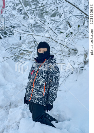 Young Boy Kneeling in Pristine Snowy Forest with Abstract Winter Jacket and Serene Expression in Wintry Landscape. Winter Wonderland Adventure: Child in Santa Hat Exploring Snowy Forest Path with 123110688
