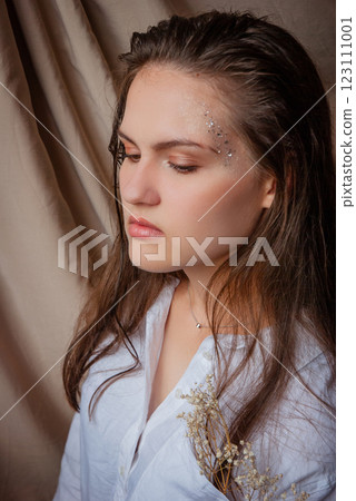 Reflective woman with sparkles and white flowers against a draped fabric background. Caucasian woman with glitter on face. Beige background Reflective woman with sparkles and white flowers against a draped fabric background. Caucasian woman with glitter on face. Beige background 123111001