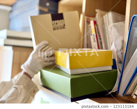 A hand taking out a scrapbook from a bookshelf with boxes and other items in front of it A hand taking out a scrapbook from a bookshelf with boxes and other items in front of it 123113184
