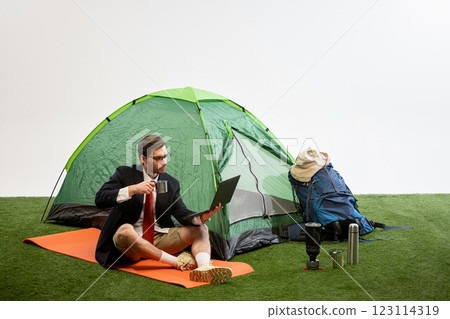 Businessman sitting on camping mat near tent typing on laptop while camping gear surrounds against white studio background. 123114319