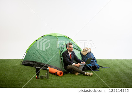 Businessman sitting near camping tent working on laptop, smiling, leaning on backpack against white studio background. Businessman sitting near camping tent working on laptop, smiling, leaning on backpack against white studio background. 123114325