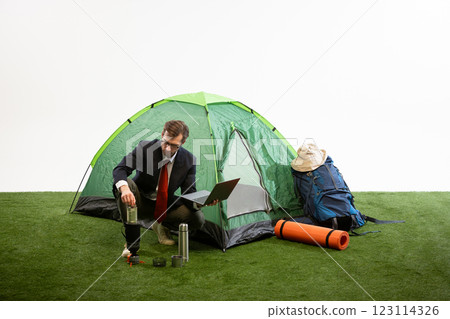 Businessman crouching near camping tent lifting coffee press while working on laptop against white studio background. 123114326