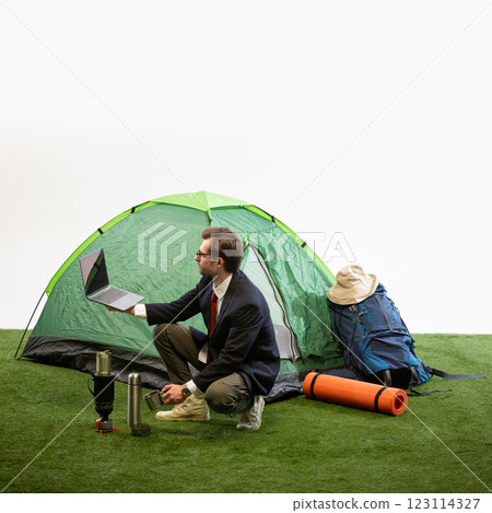 Businessman crouching near camping tent holding laptop and pouring drink from thermos against white studio background Businessman crouching near camping tent holding laptop and pouring drink from thermos against white studio background 123114327