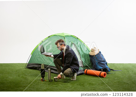 Businessman kneeling near camping tent holding laptop while preparing coffee using portable stove against white studio background. 123114328
