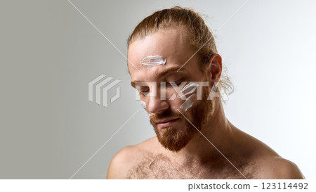 Man with tied-back hair and beard looking downward with white face cream applied, expressing calmness and skincare awareness 123114492