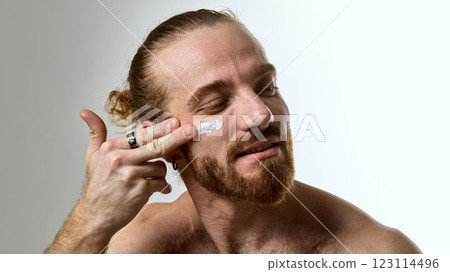 Man with tied-back hair and beard applying white face cream to cheek, showcasing smooth skin and hydration against white studio background. Man with tied-back hair and beard applying white face cream to cheek, showcasing smooth skin and hydration against white studio background. 123114496