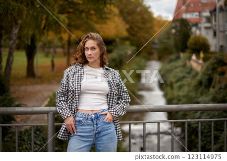 Autumn Urban Serenity: Beautiful Young Woman in Checkered Shirt and Crop Top Poses on Bridge with Vibrant Foliage and Red-Roofed Buildings. Casual Fashion Meets Nature in Tranquil Park Setting with Autumn Urban Serenity: Beautiful Young Woman in Checkered Shirt and Crop Top Poses on Bridge with Vibrant Foliage and Red-Roofed Buildings. Casual Fashion Meets Nature in Tranquil Park Setting with 123114975