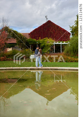 Loving Couple Embracing by a Reflective Pond in a Lush Garden with Red Ivy-Covered House and Autumn Colors 123114976
