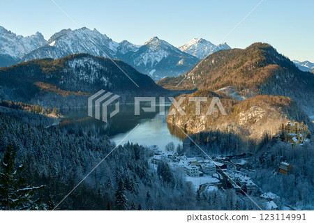 Picturesque Snowy Village by Reflective Lake in Alpine Valley with Majestic Snow-Capped Mountains and Evergreen Forest Under Clear Blue Sky Picturesque Snowy Village by Reflective Lake in Alpine Valley with Majestic Snow-Capped Mountains and Evergreen Forest Under Clear Blue Sky 123114991