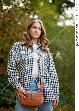 Young woman in a stylish black-and-white plaid shirt, white crop top, and blue jeans posing in a natural autumn park with a tan leather crossbody bag. 123115034