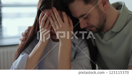 Young woman sitting on sofa, looks pessimistic and sad, almost crying, man calming her down, softly touches and hugs her. Partnership and support concept. Young woman sitting on sofa, looks pessimistic and sad, almost crying, man calming her down, softly touches and hugs her. Partnership and support concept. 123115370