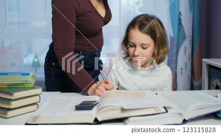 A mother helps her daughter with homework in a cozy evening room, surrounded by bookshelves encouraging learning and creativity 123115828
