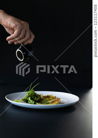 A hand pours sauce over elegantly arranged bok choy on a white plate in a dark setting. A hand pours sauce over elegantly arranged bok choy on a white plate in a dark setting. 123117408