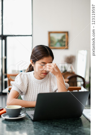 Young woman feeling stressed while working on a laptop at home, with a coffee cup on the table in a modern living room setting. 123119597