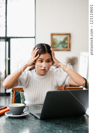 A young woman looks stressed while working on her laptop at home, with a coffee mug on the table in a bright, modern living room. A young woman looks stressed while working on her laptop at home, with a coffee mug on the table in a bright, modern living room. 123119601