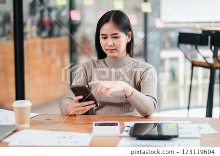 Young woman in a modern office setting, using a smartphone at a desk with digital devices and coffee. 123119604