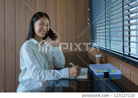 Young woman in casual white shirt smiling while talking on smartphone, working at desk with notepad and coffee 123127110
