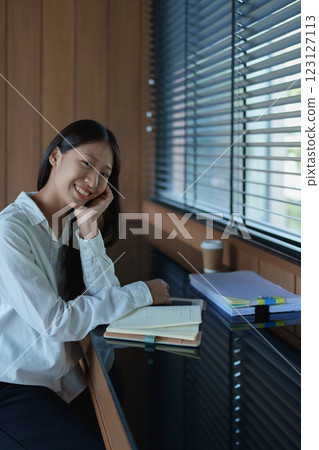 Young woman smiling while sitting at a desk with notepad, coffee, and natural light streaming through blinds 123127113