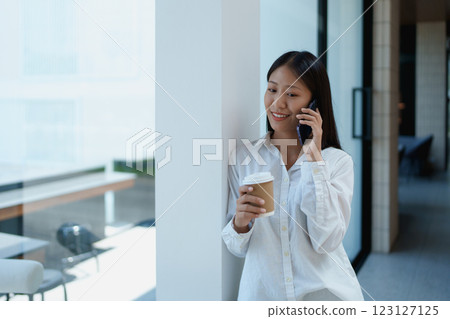 Young woman enjoying coffee while talking on a smartphone in a modern, bright office space with natural light Young woman enjoying coffee while talking on a smartphone in a modern, bright office space with natural light 123127125