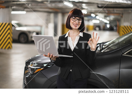 Caucasian woman in professional attire stands in parking lot holding laptop, expressing confidence with OK gesture. Portrays modern professional workplace and technology in everyday business setting. Caucasian woman in professional attire stands in parking lot holding laptop, expressing confidence with OK gesture. Portrays modern professional workplace and technology in everyday business setting. 123128511