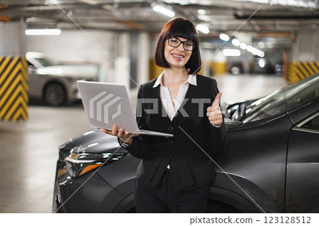Caucasian businesswoman in indoor parking lot holding laptop and showing thumbs up. Professional attire and positive attitude highlight success and confidence. Young adult female expressing. 123128512