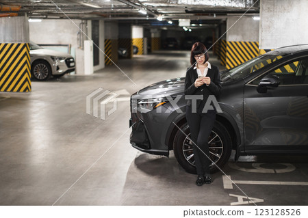 Confident young businesswoman using smartphone in modern car park. Caucasian female in business attire texting while standing next to car. Urban work life and connectivity. Confident young businesswoman using smartphone in modern car park. Caucasian female in business attire texting while standing next to car. Urban work life and connectivity. 123128526