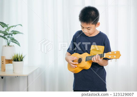A young boy in a blue shirt plays yellow ukulele in living room. The white curtains in the background add a light, airy touch to cheerful moment, highlighting his playful expression and musical joy. 123129007