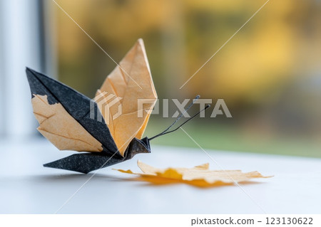 A detailed paper butterfly, folded from brown and black sheets, sits gracefully on a white surface. The background features soft, blurred autumn colors, enhancing the tranquility of the moment 123130622