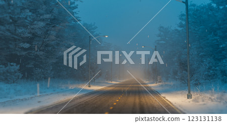 Winter road with snow covered trees on both sides, illuminated by streetlights at dusk, creating serene and peaceful atmosphere in forest 123131138