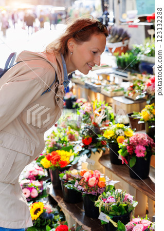 Young adult joyful woman kneels to choose buy colorful potted flowers at outdoor market stall. Sale of blooming plants in pots at botanical street German store on sunny spring day 123132288