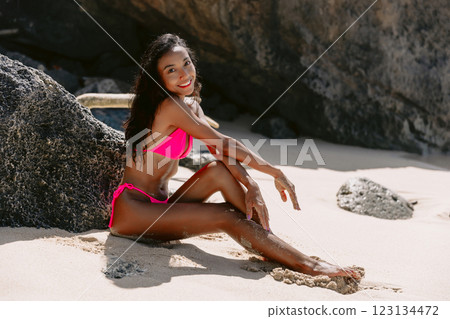 Smiling woman in a bikini sitting on sandy sunny beach, embracing summer vibes 123134472