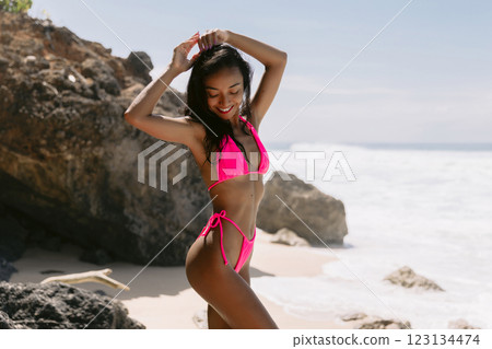 Smiling woman in a bikini enjoying a sunny beach day, embracing summer vibes with ocean waves and rocky cliffs behind her 123134474