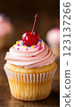 Vanilla cupcake with deep pink frosting and glossy red cherry, moody lighting enhancing golden cake crumb, placed on dark textured wooden backdrop with scattered colorful sprinkles, blurred background 123137266