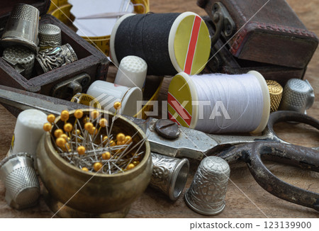 Sewing needles stuck in a white paper, Thimbles in small wooden treasure chest, Spools of black and white thread, Steel scissors and Measuring tape including pins in brass cup on wooden table. 123139900