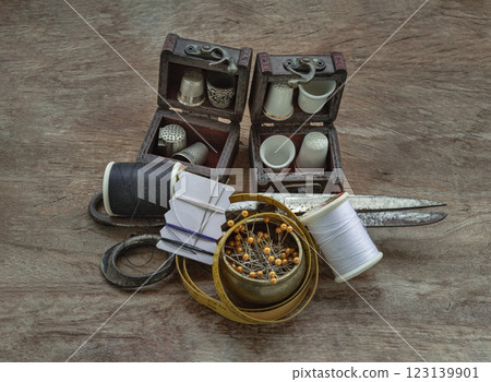 Thimbles in small wooden treasure chest, Sewing needles stuck in a white paper, Spools of black and white thread, Steel scissors and Measuring tape including pins in brass cup on wooden background. Thimbles in small wooden treasure chest, Sewing needles stuck in a white paper, Spools of black and white thread, Steel scissors and Measuring tape including pins in brass cup on wooden background. 123139901