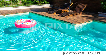 Turquoise swimming pool with pink inflatable ring, wooden deck, and modern lounge chairs in sunny backyard 123141915