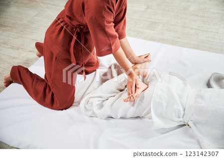 Masseuse making thai yoga massage. Woman therapist doing traditional massage treatment, stretching female patient's body. Client lying on mat, enjoying hand massage in calm atmosphere. Cropped view. 123142307