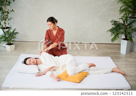 Masseuse making thai yoga massage. Woman therapist doing traditional massage treatment for female patient indoors. Client lying on mat, meditating, enjoying hand massage in calm atmosphere. 123142311