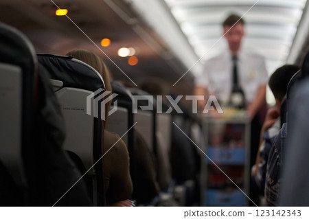 A male steward who distributes food and drinks on board the aircraft. The passengers are sitting in their seats and watching. View along the row. High quality photo A male steward who distributes food and drinks on board the aircraft. The passengers are sitting in their seats and watching. View along the row. High quality photo 123142343