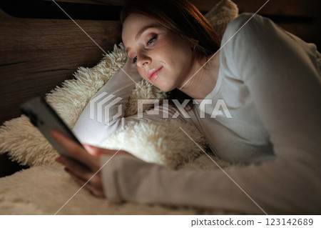 Peaceful redhead woman lying on bed, using smartphone in dark bedroom at night, enjoying leisure time while smiling looking to illuminated screen, radiating happiness. 123142689