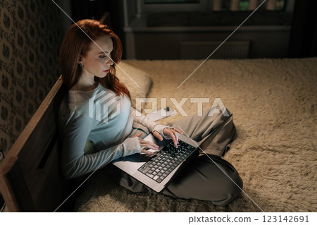 Top view of focused female freelancer working late on laptop sitting in bed, face illuminated by screen glow in bedroom, demonstrating dedication to tasks. Successful woman discussing new project Top view of focused female freelancer working late on laptop sitting in bed, face illuminated by screen glow in bedroom, demonstrating dedication to tasks. Successful woman discussing new project 123142691