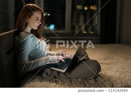 Successful businesswoman working late on laptop in bed, illuminated by screen light, demonstrating dedication and work-life balance in home office setting. Female freelancer typing on keyboard. 123142695