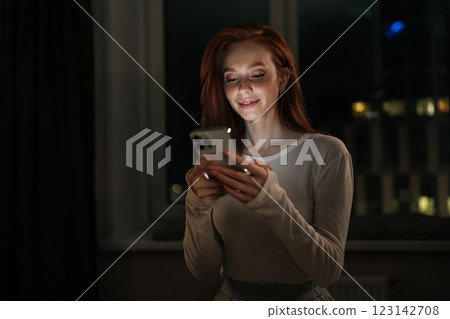 Attractive happy young woman with long red hair smiling using mobile phone at night, sitting comfortably by window in dark living room, happily typing message and connecting online, looking to screen. 123142708