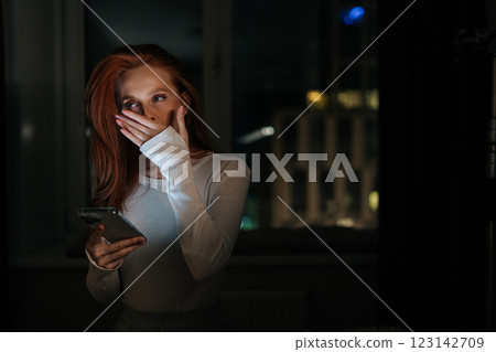 Portrait of tired young woman yawning and covering mouth with hand browsing internet while chatting in social media at night sitting on bed by window with blurred bokeh city lights, in dark bedroom. Portrait of tired young woman yawning and covering mouth with hand browsing internet while chatting in social media at night sitting on bed by window with blurred bokeh city lights, in dark bedroom. 123142709