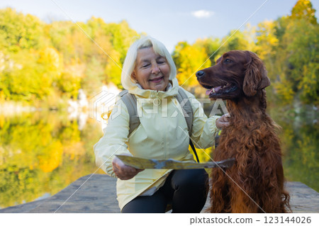 Happy senior 60s woman holds travel map searching location, with her best friend irish setter dog wear casual clothes in the autumn forest. Happy senior 60s woman holds travel map searching location, with her best friend irish setter dog wear casual clothes in the autumn forest. 123144026