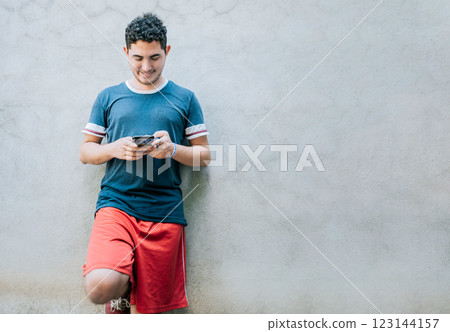 Happy guy leaning on a wall using smartphone. Smiling young man leaning on a wall sending a text message Happy guy leaning on a wall using smartphone. Smiling young man leaning on a wall sending a text message 123144157
