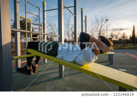 young athletic man exercising crunches on bench at fitness calisthenics workout at street gym during sunset, body improvement and strength training 123146819