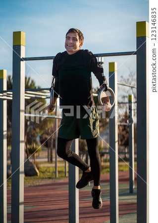 young athletic man exercising rings at fitness calisthenics workout and making pull-ups at street gym during sunset, body improvement and strength training concept young athletic man exercising rings at fitness calisthenics workout and making pull-ups at street gym during sunset, body improvement and strength training concept 123146835