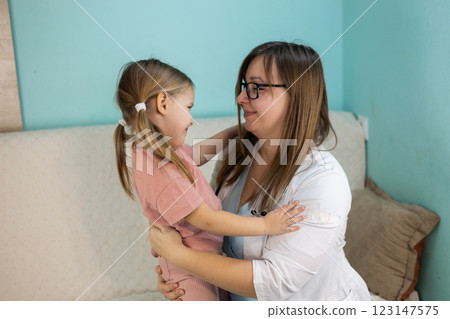 Compassionate doctor in white coat examines little girl in cozy home environment. Child smiles, feeling calmed by doctor's gentle presence and attentive care. Concept medicine and health care. 123147575