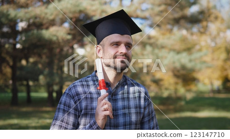 A joyful man dressed in graduation attire is holding his diploma proudly outdoors amidst the vibrant autumn foliage 123147710